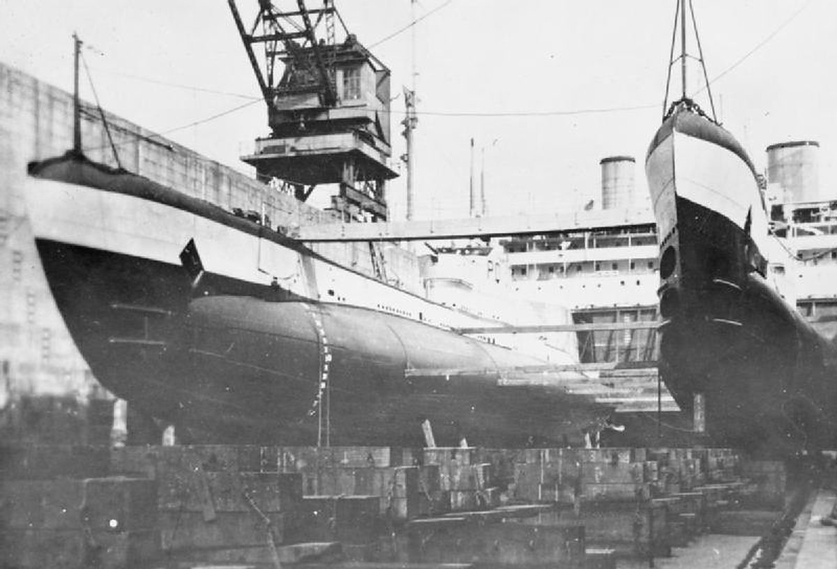 Siyah beyaz fotoğraf: kuru havuzda takozlar üzerinde duran iki denizaltı, solda uzun gövdesi görünen tekne, sağda burnu havaya kalkık ikinci tekne, arkada vinç ve yolcu gemisi bacaları