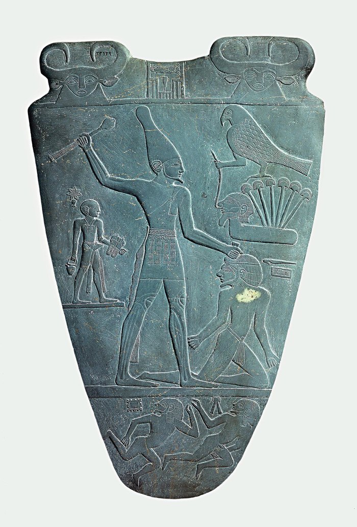 Narmer Paleti'nin ön yüzü: beyaz taçlı kral Narmer, sağ elinde kaldırdığı topuzla önünde diz çökmüş bir adamın başına vurmak üzere; alt şeritte yere serilmiş iki figür