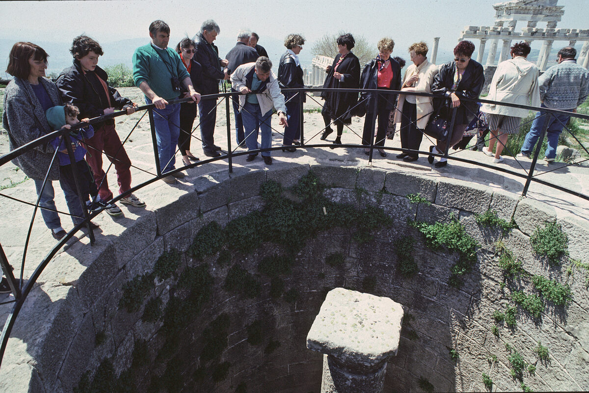 Bergama akropolünde taş örgülü yuvarlak bir sarnıcın ağzı; korkuluğun çevresinde aşağı bakan ziyaretçiler, sarnıcın ortasında tek bir sütun başlığı
