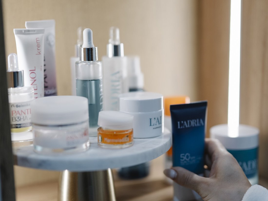 Bir rafta yan yana duran serum, krem ve güneş kremi şişeleri, bir el güneş kremini alıyor
