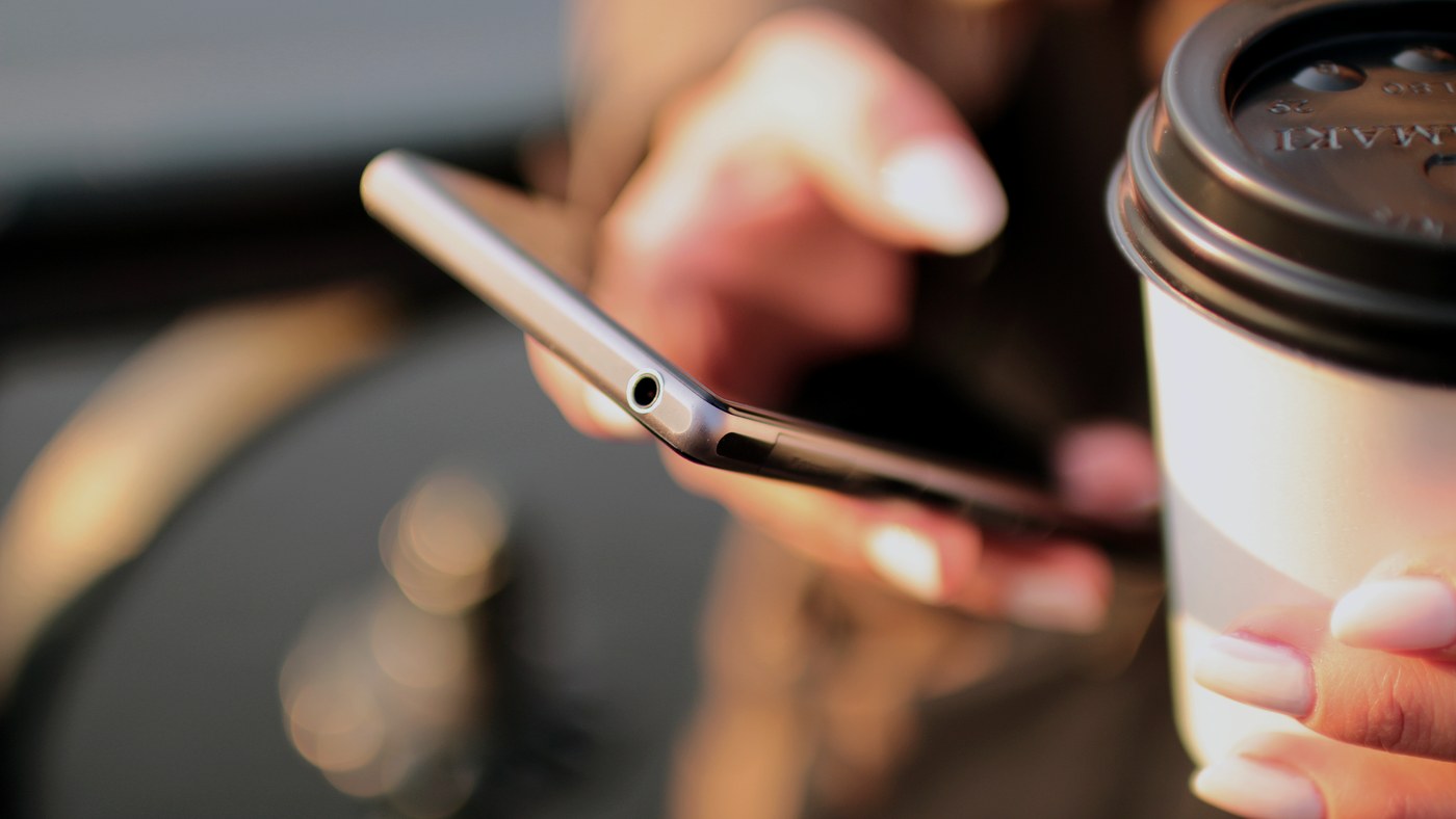 Bir elde kahve, diğerinde akıllı telefon tutan eller