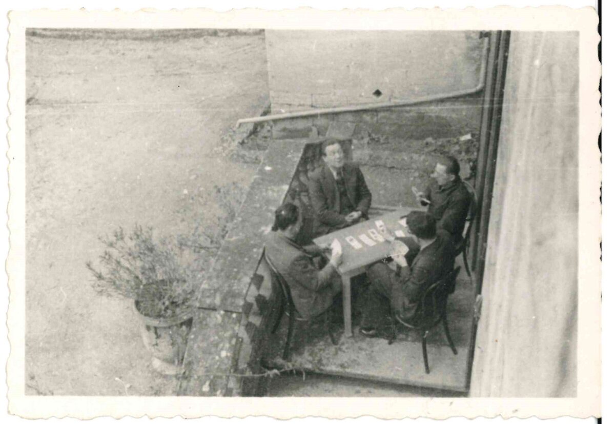 Siyah beyaz eski fotoğraf: bir avluda küçük bir masanın çevresinde oturmuş, iskambil oynayan dört adam, yukarıdan çekilmiş
