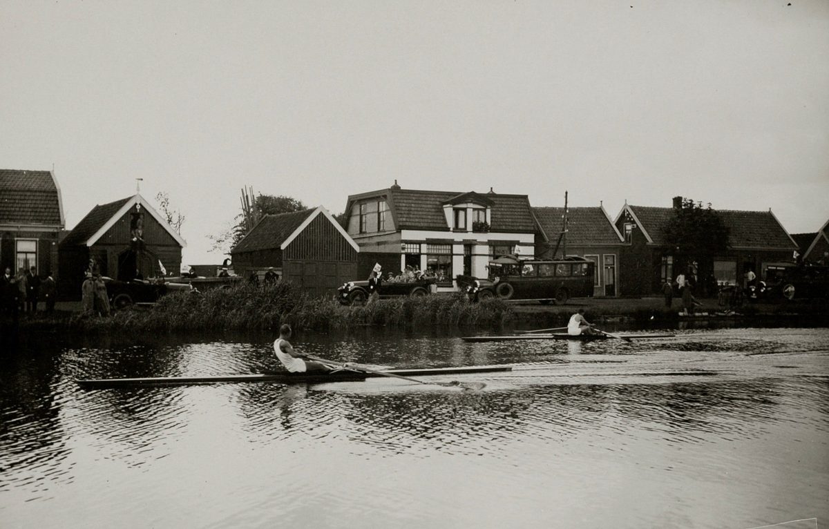 1928 Amsterdam Olimpiyatları'nda Ringvaart kanalında iki tek kişilik kürek teknesi yarışıyor; kıyıda ahşap evler, otomobiller ve seyirciler