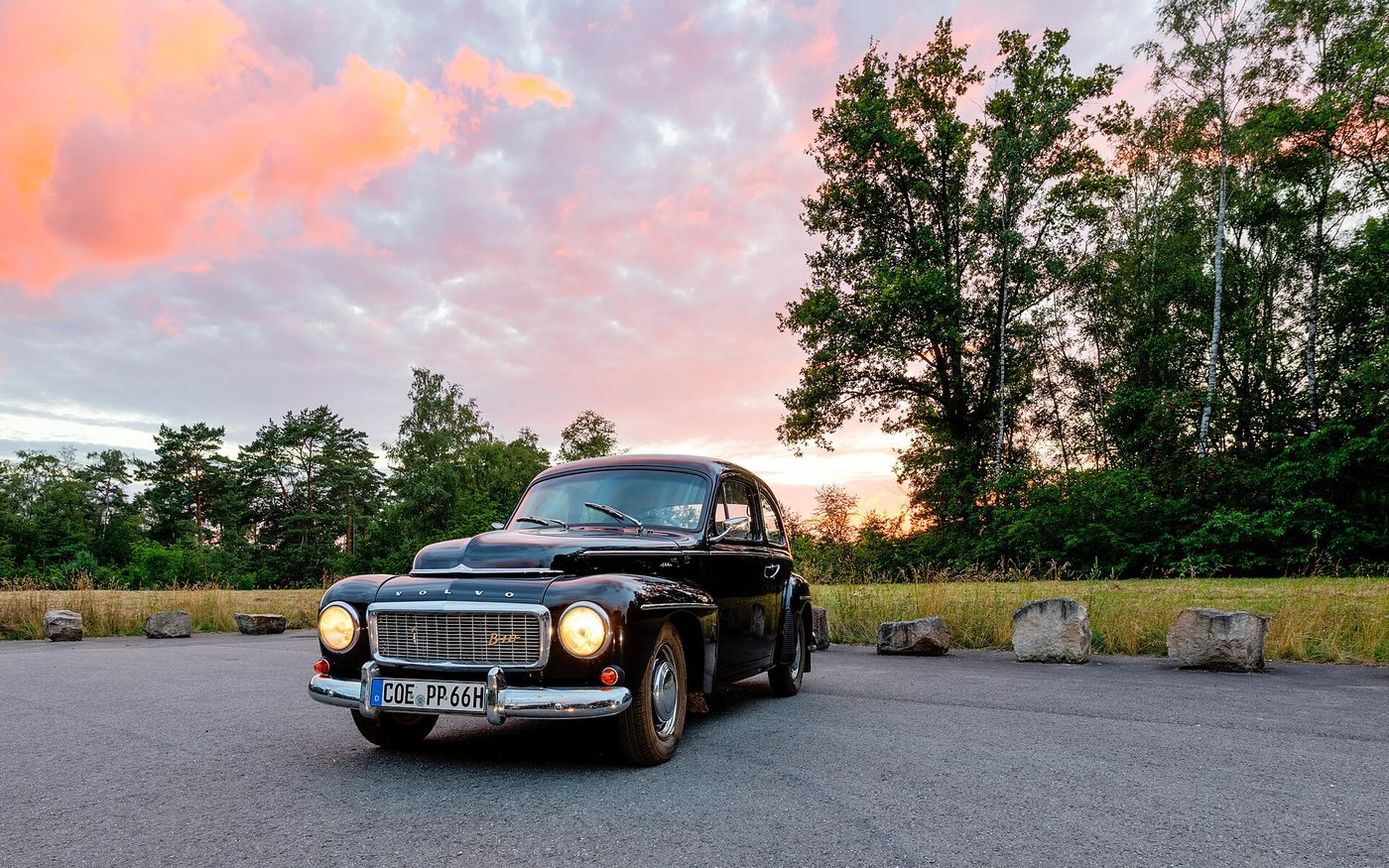 Siyah bir 1961 model Volvo PV544 akşam gökyüzü altında, ağaçlarla çevrili bir otoparkta duruyor