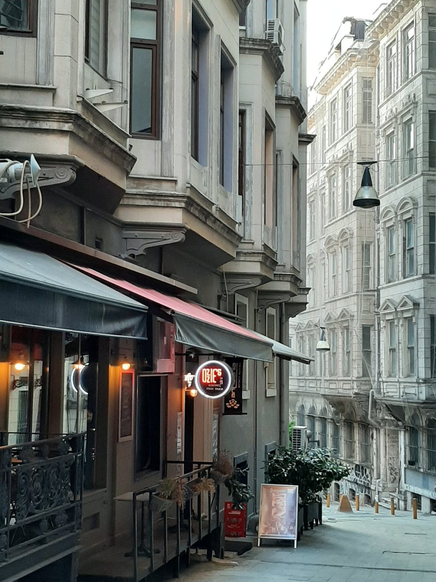 Asmalımescit’te dar bir yokuş sokak, solda tenteli bir kafe ve cumbalı eski binalar, sağda yüksek taş cepheli apartmanlar
