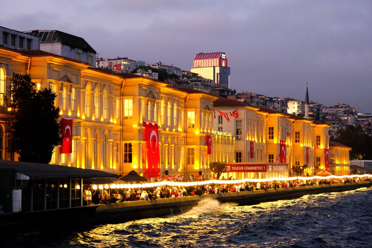 Boğaz kıyısında, akşam ışıklarıyla aydınlanmış sarı cepheli uzun saray binaları, cephelerde Türk bayrakları, önlerinde rıhtımda kalabalık