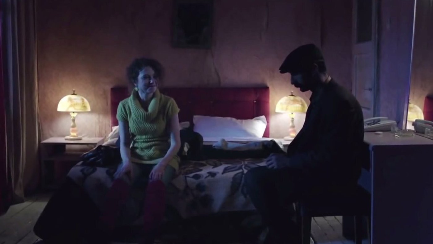 Soğuk filminden bir kare: loş, abajurlu bir otel odasında yeşil kazaklı genç bir kadın yatağın kenarında, kasketli bir adam ise sırtı dönük oturuyor