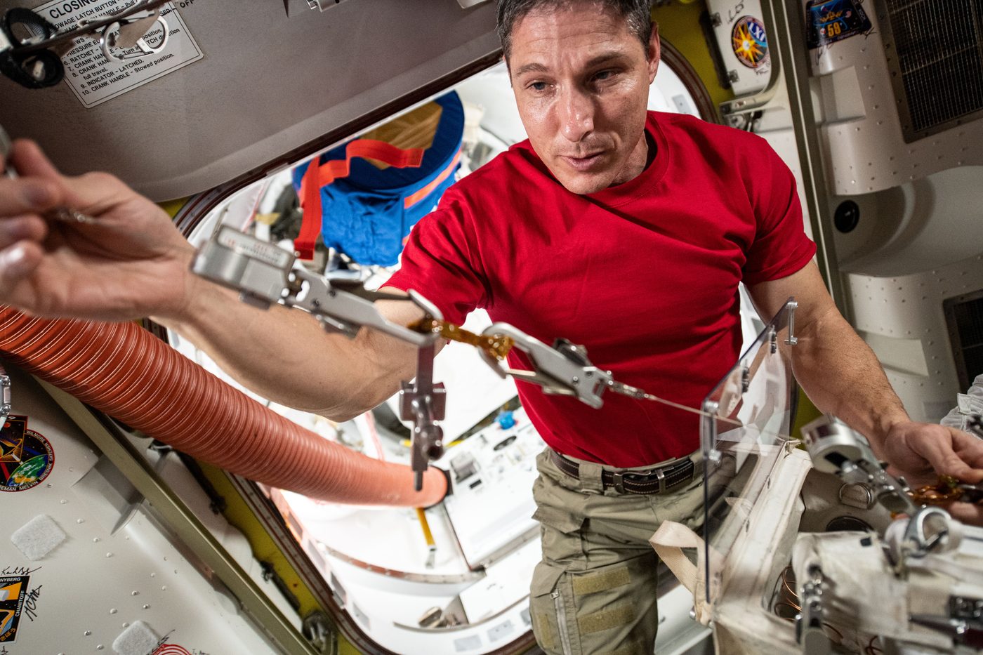 Kırmızı tişörtlü astronot Michael Hopkins istasyonun içinde metal kancalı emniyet iplerini elden geçiriyor