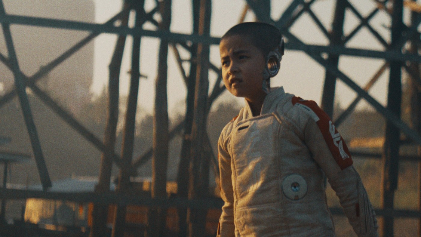 Yaratıcı filminden bir kare: kafasının yanında metal bir aparat olan küçük Alphie, ahşap iskeleli bir köprünün üstünde duruyor